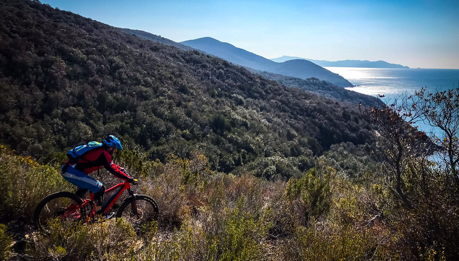 Un biker durante un'escursione in e-MTB enduro su un sentiero singletrack nella macchia mediterranea, con vista panoramica sul mare e sulle colline della Maremma toscana.