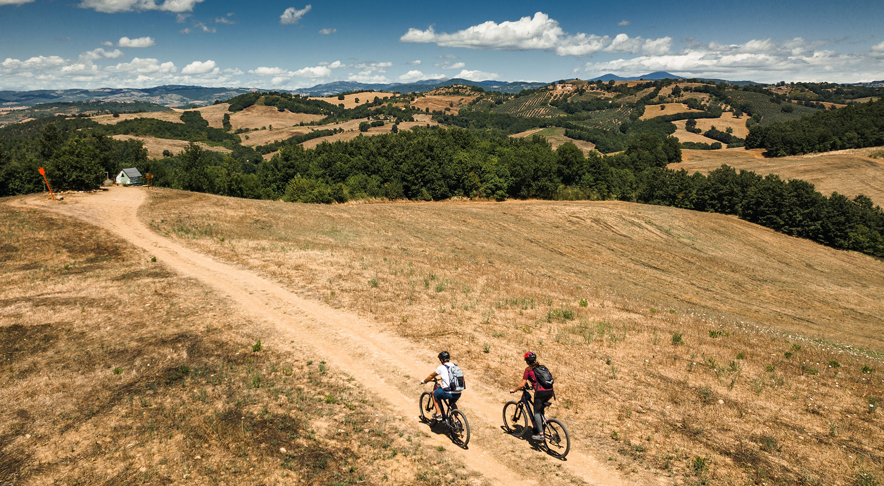 Veduta aerea di due ciclisti in e-bike che percorrono un sentiero sterrato panoramico sulle colline della Fattoria La Maliosa in Maremma.
