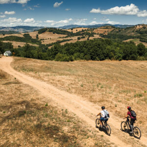 Veduta aerea di due ciclisti in e-bike che percorrono un sentiero sterrato panoramico sulle colline della Fattoria La Maliosa in Maremma.